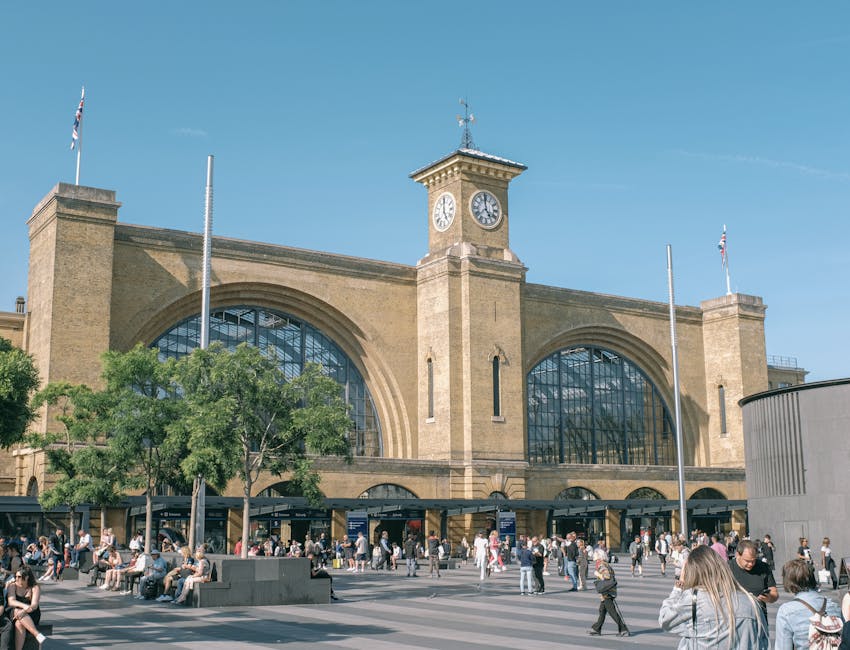 A large historic train station with a prominent central clock tower displaying the time at approximately 2:55, constructed from tan brick with arched glass windows. The building features multiple entrances at ground level, with a crowd of pedestrians walking on the paved pavement in front of it. Some individuals are seated on concrete benches, while others are standing or walking. A tall flagpole with the Union Jack flag is positioned on the left side of the station, and another flagpole without a flag is on the right. Trees with green foliage partially obscure the lower part of the station. The scene is set during daylight with a clear blue sky, and the overall environment suggests busy urban activity typical around a central transportation hub. This setting aligns with the kind of area where Man With a Van Earls Court provides relocation and furniture transport services, supporting home removals and packing tasks.