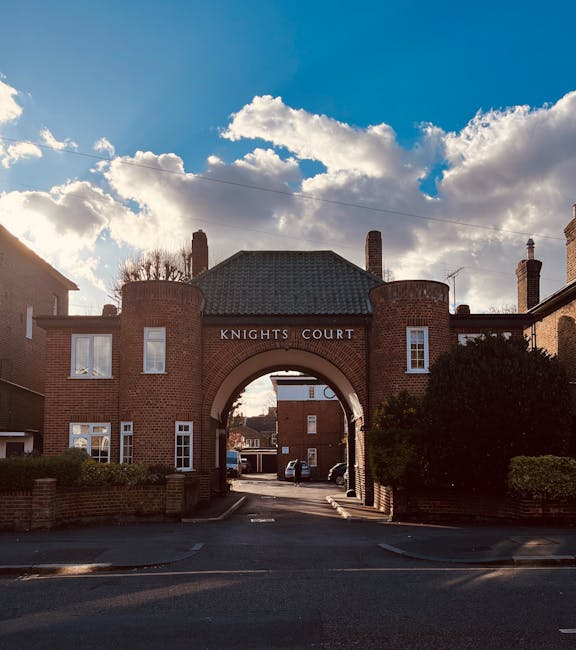 A large brick residential building with an arched entrance labeled 'Knights Court' under a blue sky with scattered white clouds. The building features multiple chimneys, white-framed windows, and neatly trimmed greenery along the sides. In the foreground, a paved driveway leads through the archway into the internal courtyard, where several vehicles are parked. Outside the entrance, a moving company, Man With a Van Earls Court, is seen actively involved in home relocation logistics, with visible cardboard boxes, wrapped furniture, and packing materials being loaded onto a van. The scene captures the loading process inside the property, with some furniture pieces and boxes being carried by movers, demonstrating furniture transport and packing during a house removal. The image emphasizes professional moving practices, including the careful handling of household items in a typical residential area of Old Brompton Road, supporting services offered by [COMPANY_NAME].
