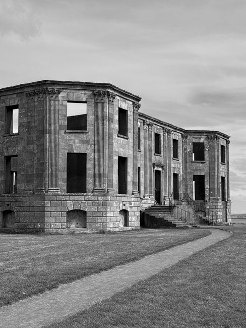 A black and white photograph of a historic, semi-circular building with large, empty window openings and decorative vertical columns on its façade, situated on a grassy area with a winding concrete pathway leading up to the entrance stairs. The building appears to be an old, possibly abandoned or unfinished structure, with visible weathering on the stone exterior. The surrounding environment is open with a cloudy sky overhead. This image, associated with house removals and furniture transport services provided by Man With a Van Earls Court for home relocation or moving projects on Old Brompton Road, captures the architectural setting that could be part of a larger property undergoing packing and moving activities.